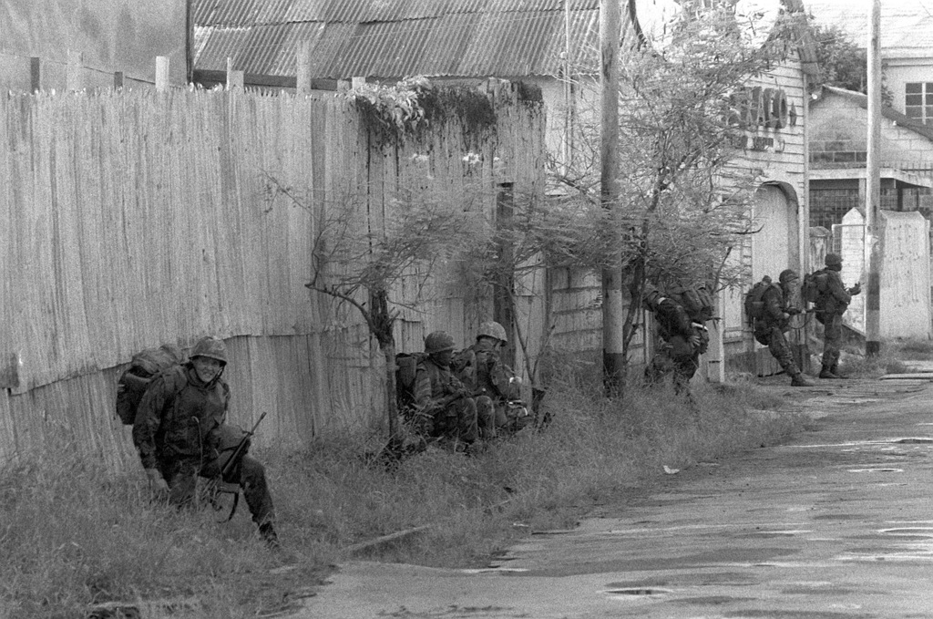 Marines, armed with M16A1 rifles, stand along a wall outside a gas ...