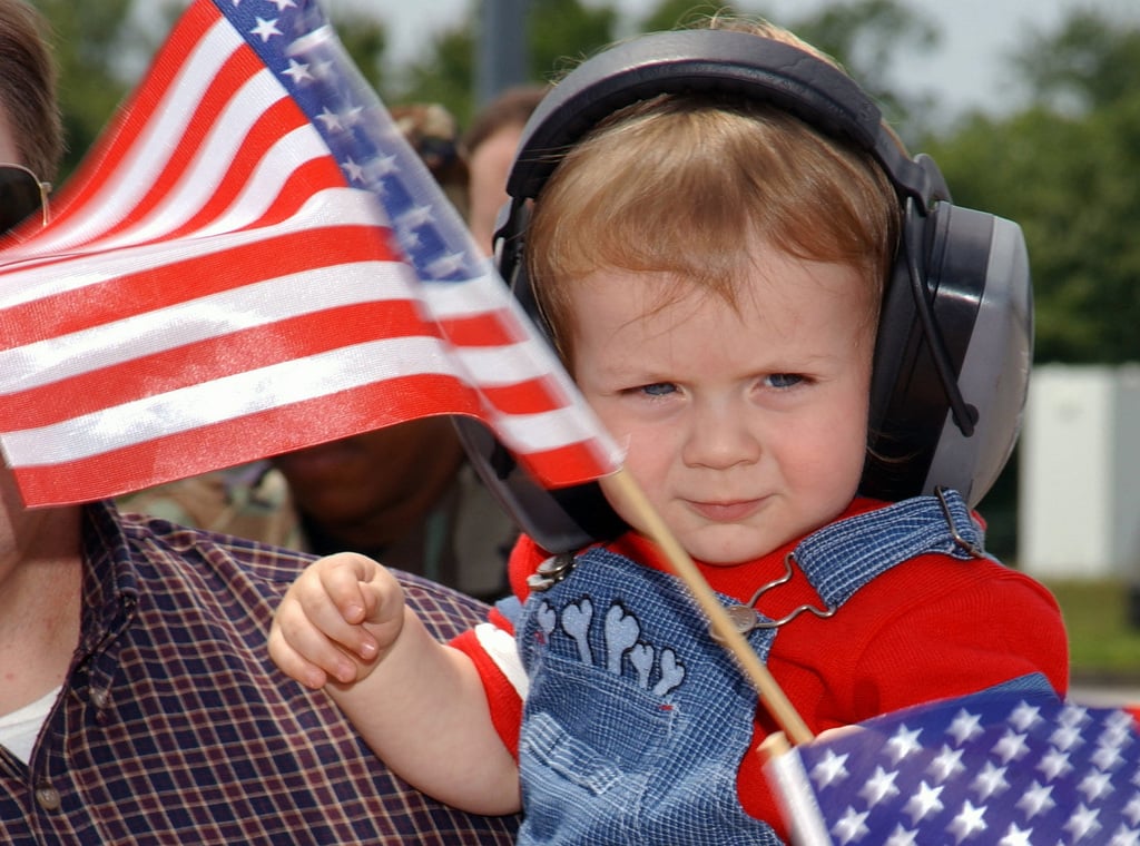 A young child waving an American Flag and wearing ear muffs awaits the ...